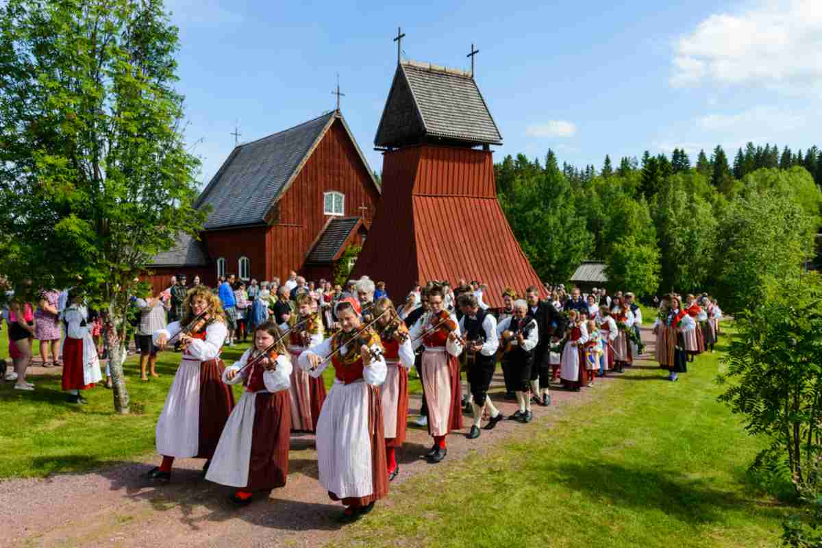 Eine Gruppe von Musikern in typischer schwedischer Tracht, angeordnet in Reihen zu je drei neben einer roten Kirche, während sie zur Ehre des Sommerfestes Mittsommer Geige spielen.
