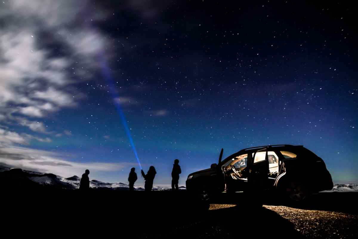 Eine Gruppe von Menschen, die im Schatten unter einem sternenübersäten Himmel steht, beleuchtet den Himmel mit einer Taschenlampe, während neben dem Auto geparkt wird.