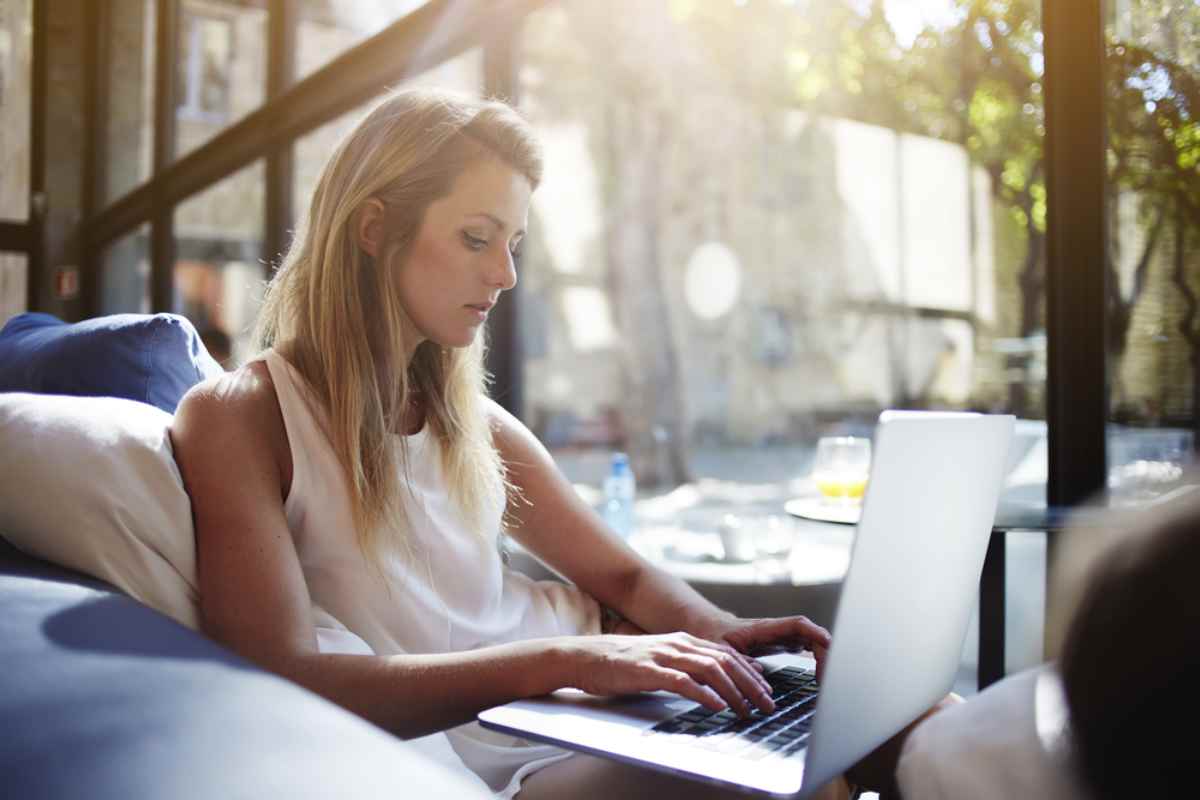 Eine blonde Frau in einem, wie es scheint, Kaffeehaus surft im Internet mit ihrem Computer neben einer Tasse Kaffee.