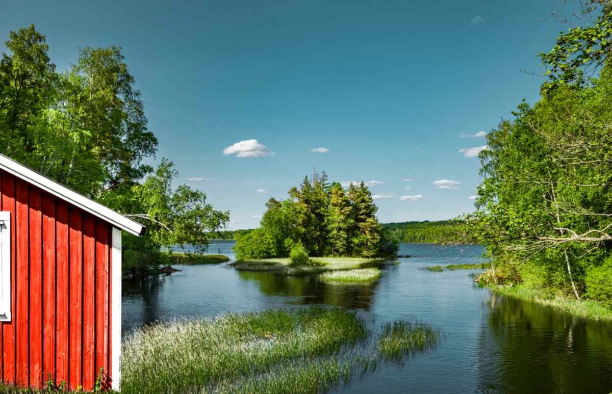 A lake with trees in and around it stretches next to what appears to be a camping cabin in Sweden on a sunny day.