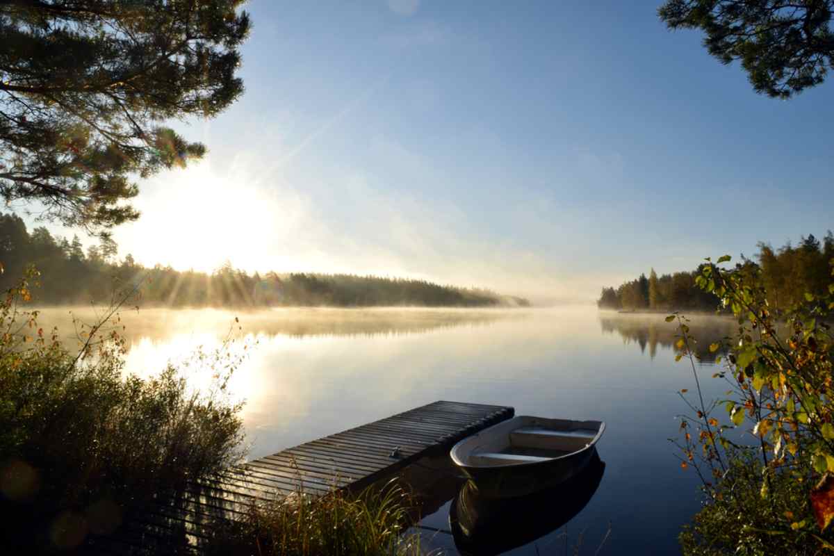 Dimmig soluppgång över en stilla skogssjö med liten träbrygga och roddbåt, en fridfull morgonscen längs en husbilsrutt i Småland.