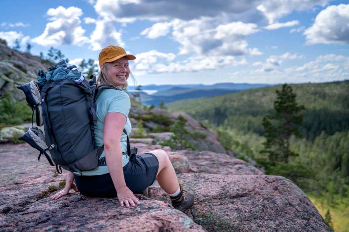 Leende vandrare med stor ryggsäck sitter på rosa klippa med skogsklädda kullar och blå himmel bakom, ett typiskt vandringsstopp på en roadtrip i södra Sverige.