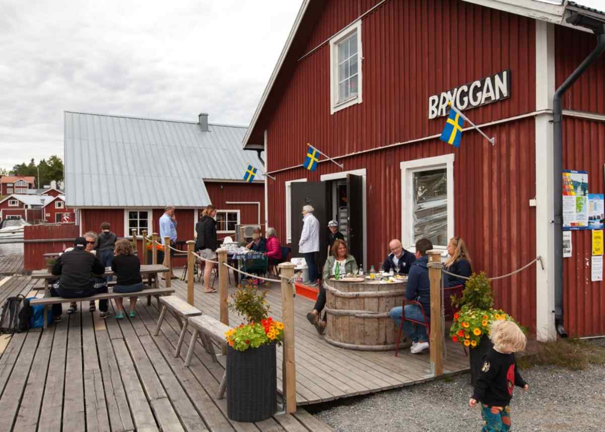 Människor som njuter av fika utanför café Bryggan i Bönhamn, sittande på en träbrygga i hamnen kantad av röda stugor och svenska flaggor.
