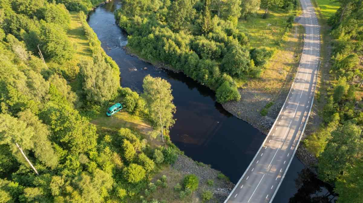 Aerial view of a campervan parked by a river beside a bridge, surrounded by forest, a scenic break on a Stockholm road trip.