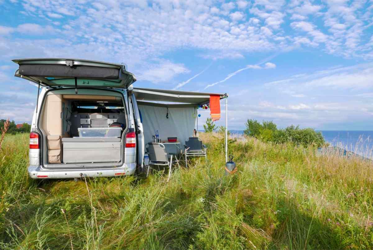 Campervan parked on a grassy coastal meadow with the rear hatch open and an awning set up with chairs and a table by the sea.