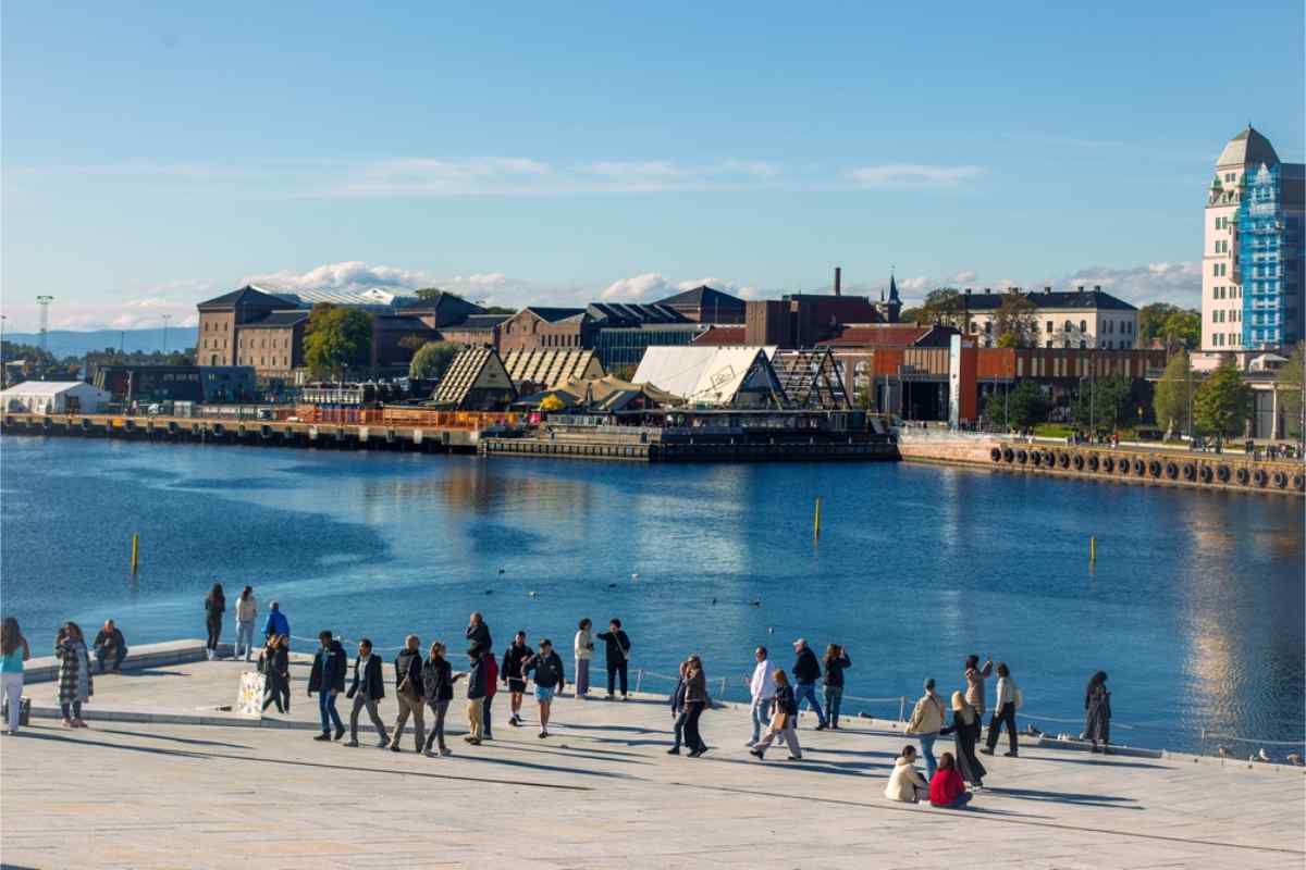 People walking and relaxing by a modern waterfront promenade with city buildings and a calm harbor on a sunny day.