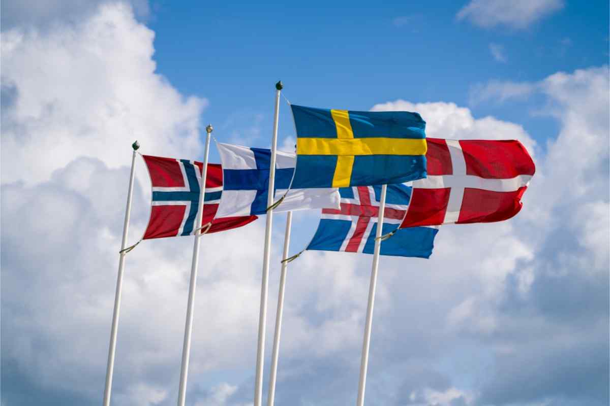 Nordic flags of Norway, Finland, Sweden, Iceland, and Denmark flying on tall poles against a bright, partly cloudy sky.